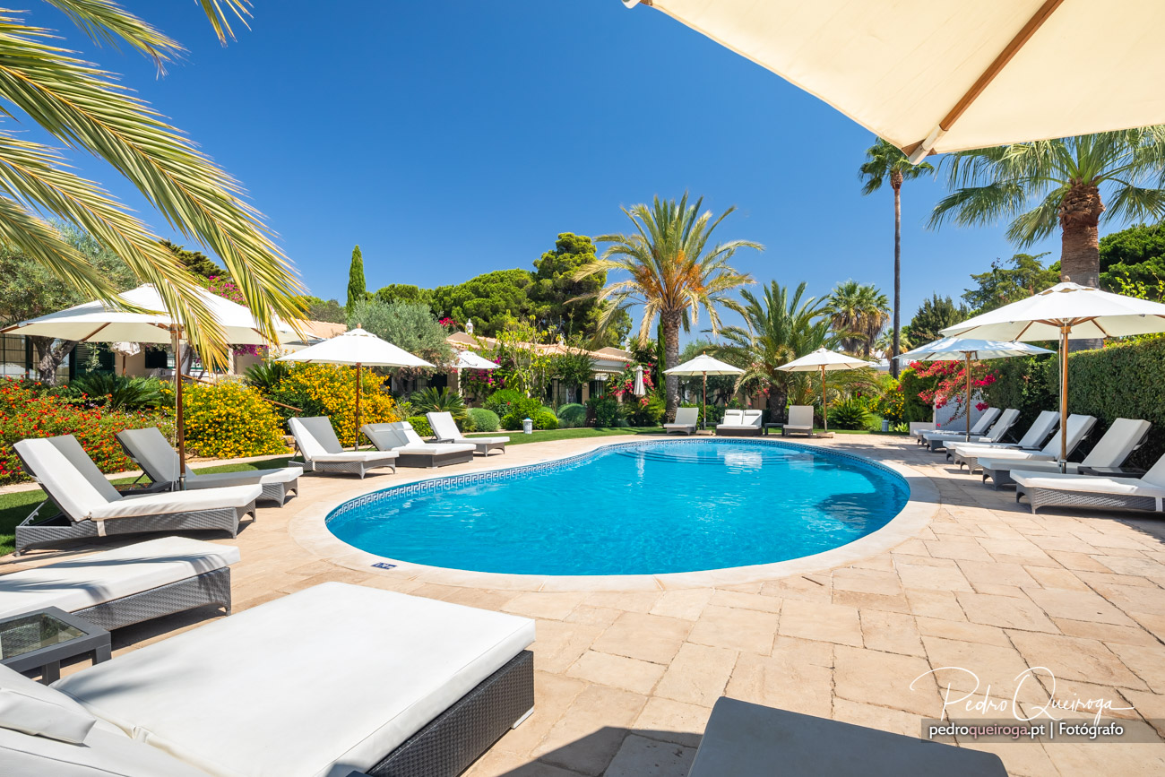 Piscina azul rodeada de espreguiçadeiras, chapéus-de-sol brancos, palmeiras e jardim florido sob céu luminoso.