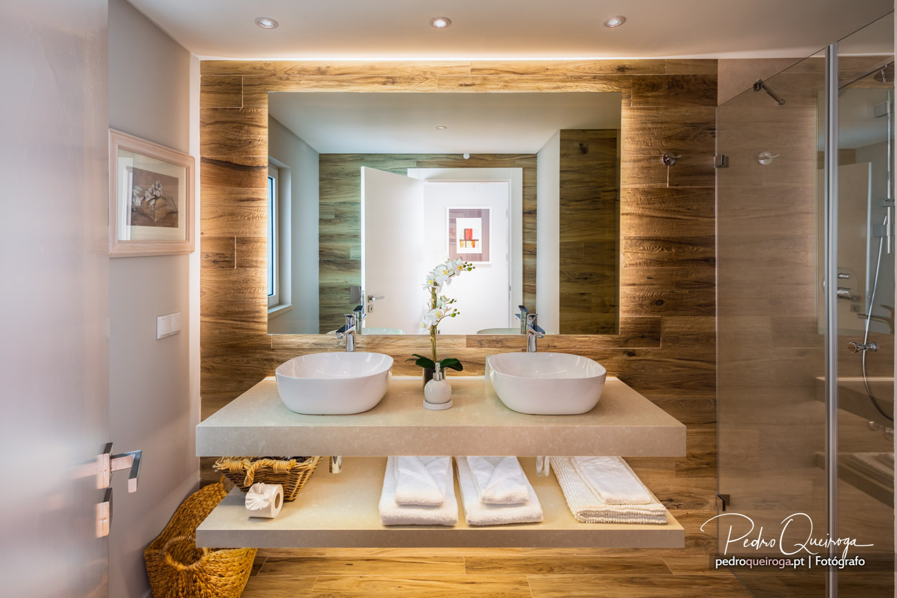 Casa de banho moderna com duplo lavatório sobre bancada bege, revestimento em madeira e espelho retroiluminado central.