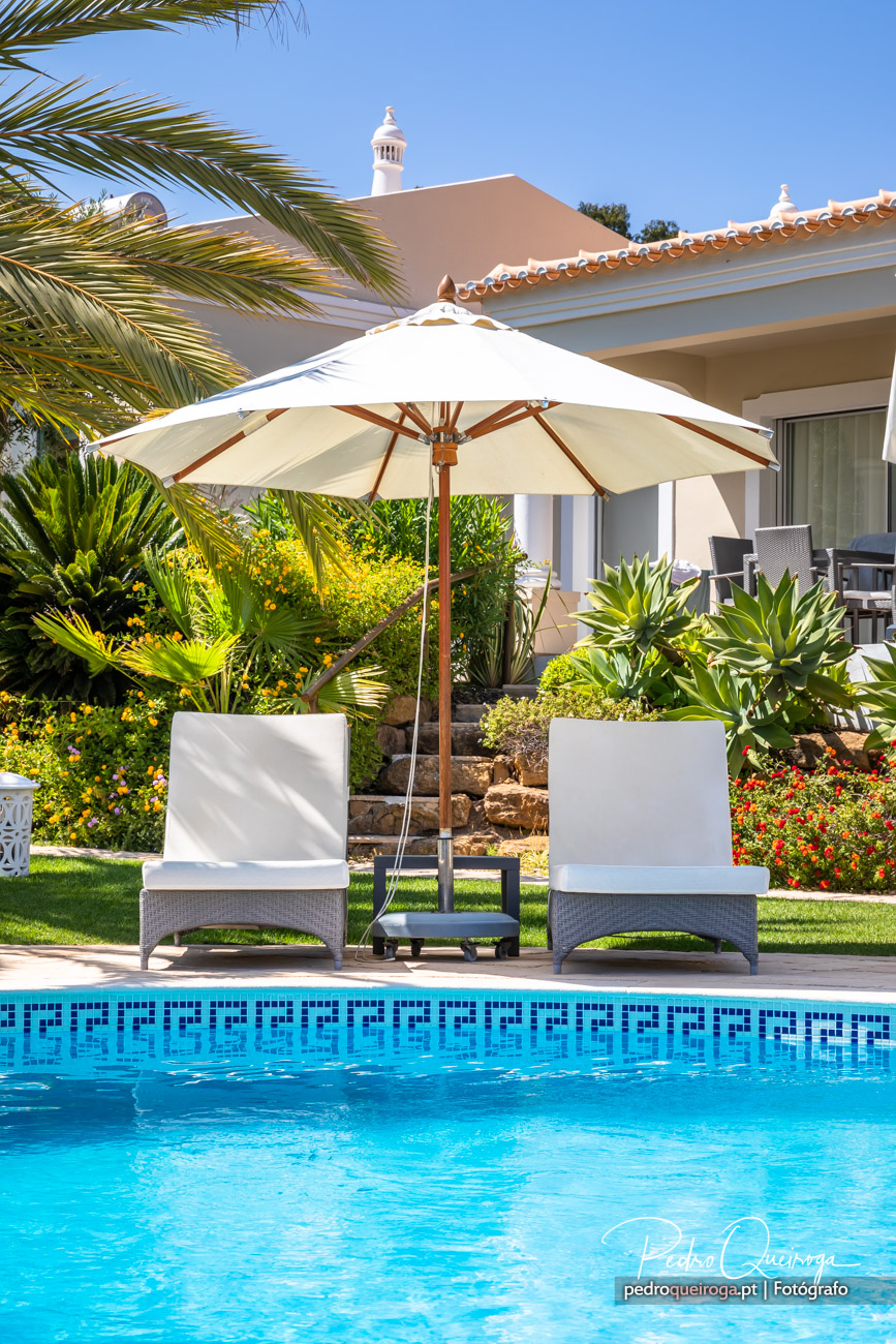 Piscina azul cristalina com espreguiçadeiras e chapéu de sol, rodeada de jardim tropical e villa algarvia ao fundo.