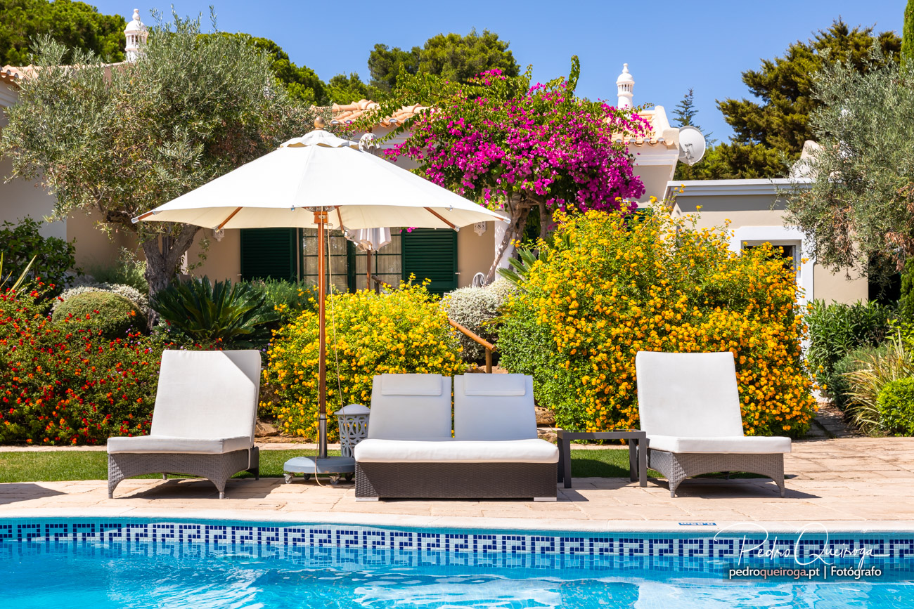 Piscina com espreguiçadeiras brancas e chapéu de sol em frente a moradia com jardim florido colorido