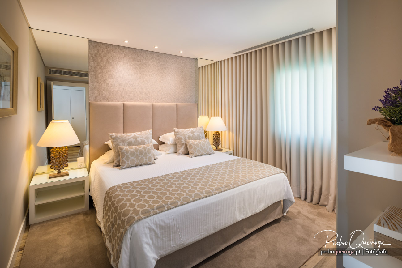 Quarto elegante com tons beige e branco, cabeceira almofadada, cortinados verticais e iluminação ambiente suave