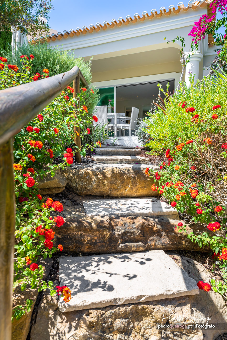 Escadaria em pedra natural rodeada de flores vermelhas e laranja, conduz a terraço com telhado de telha tradicional