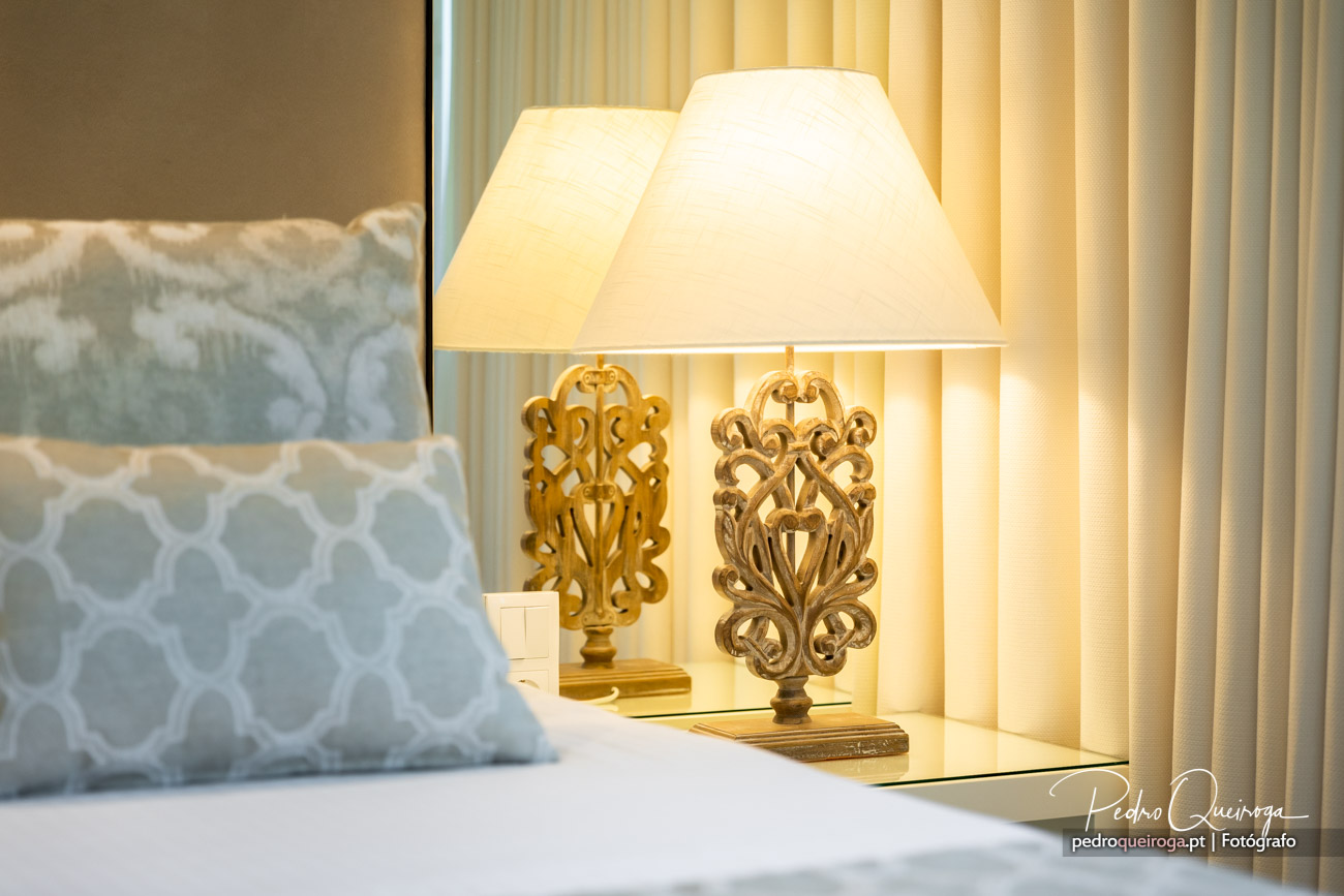 Candeeiro de mesa com base ornamentada dourada e abajur creme, junto a almofada azul com padrão geométrico branco.