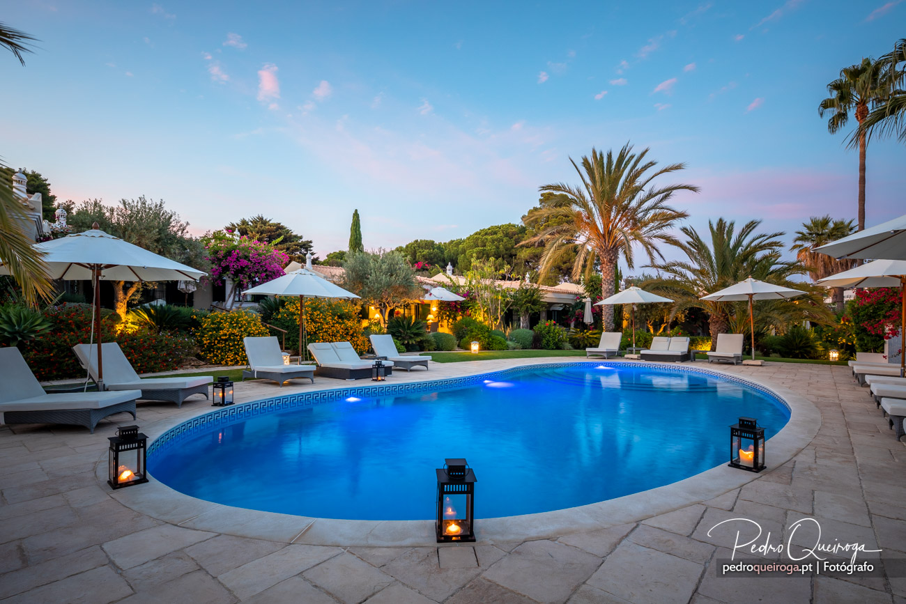 Piscina iluminada ao entardecer rodeada por espreguiçadeiras, palmeiras e jardim exuberante num ambiente tropical sereno.