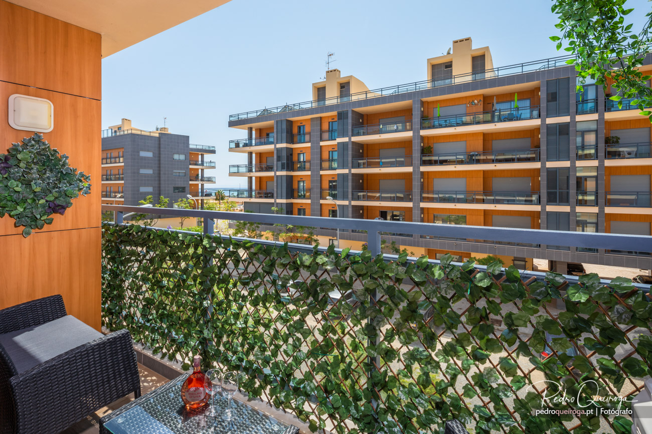 Varanda com plantas trepadeiras e mobiliário em fibra preta, com vista para edifícios modernos de tons laranja e cinza.