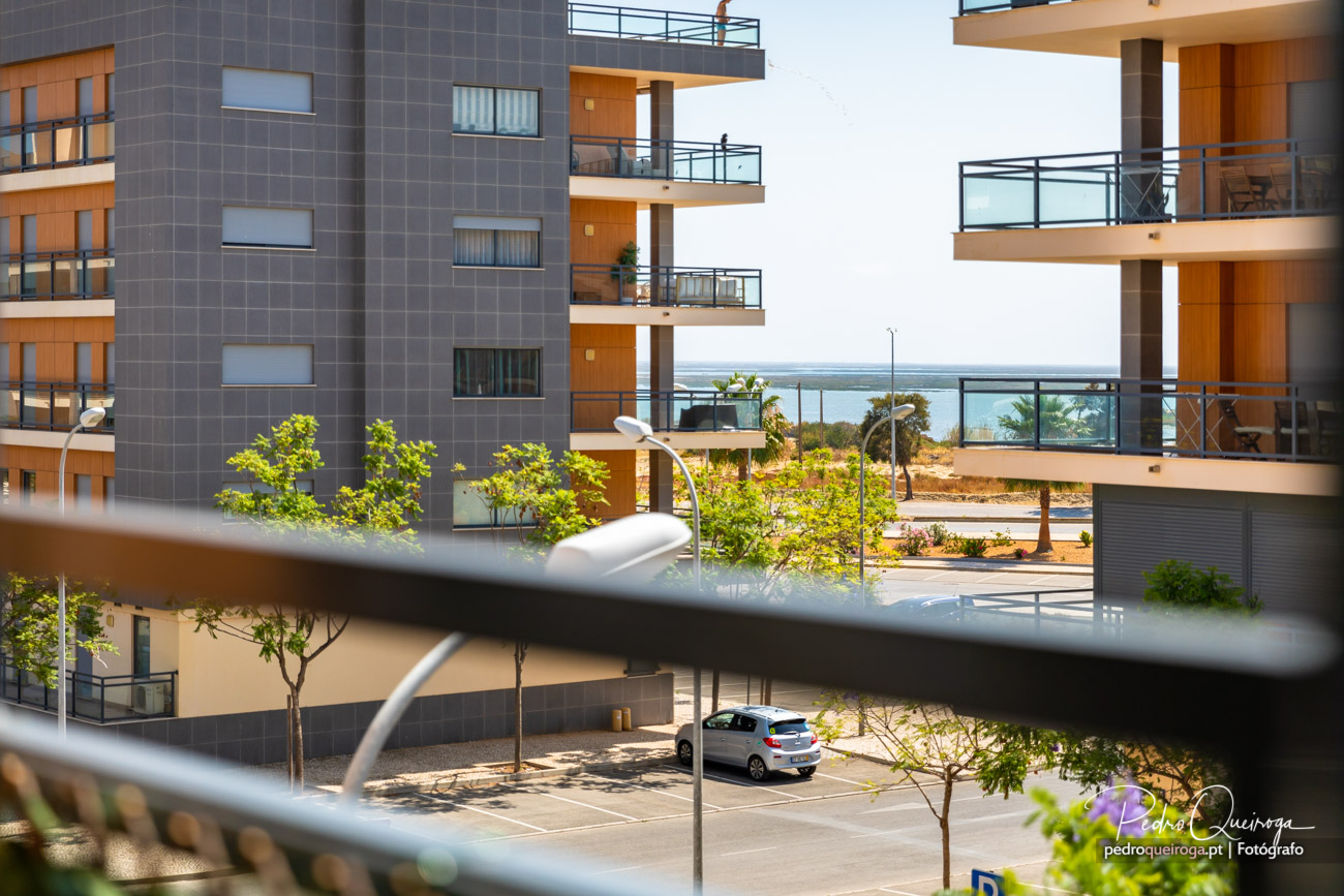 Vista através de varanda com edifícios modernos, varandas amplas, árvores jovens e mar ao fundo num dia solarengo