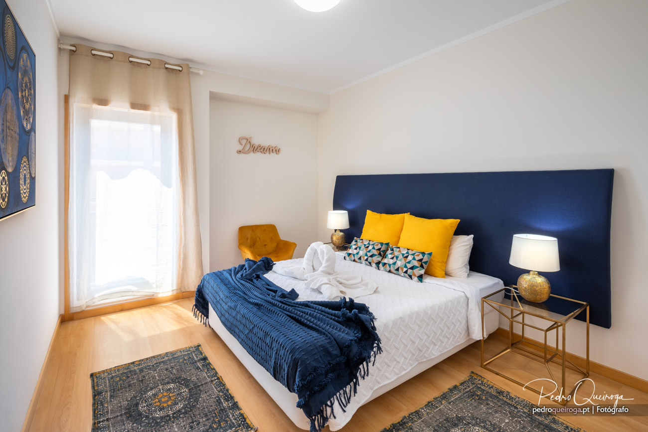 Quarto luminoso com cabeceira azul-marinho, almofadas amarelas e manta azul sobre pavimento em madeira clara.