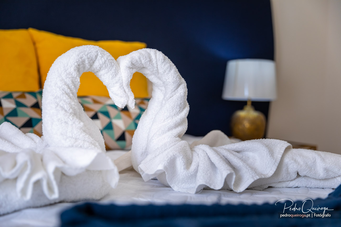 Toalhas dobradas em forma de cisnes sobre a cama, com almofadas amarelas e decoração azul ao fundo.