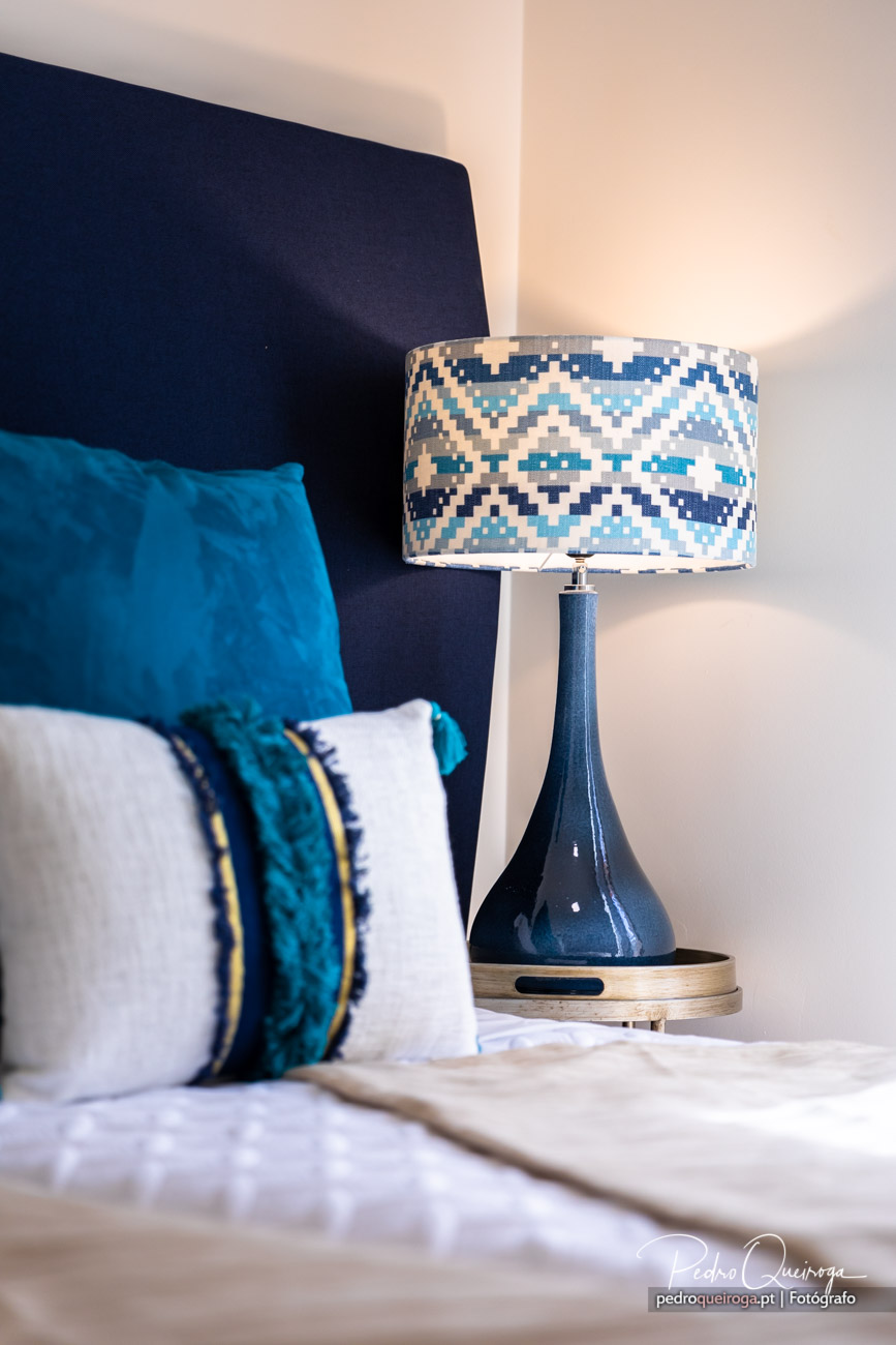 Candeeiro de mesa com padrão geométrico azul junto a cama com almofadas, detalhe de arquitetura de interiores