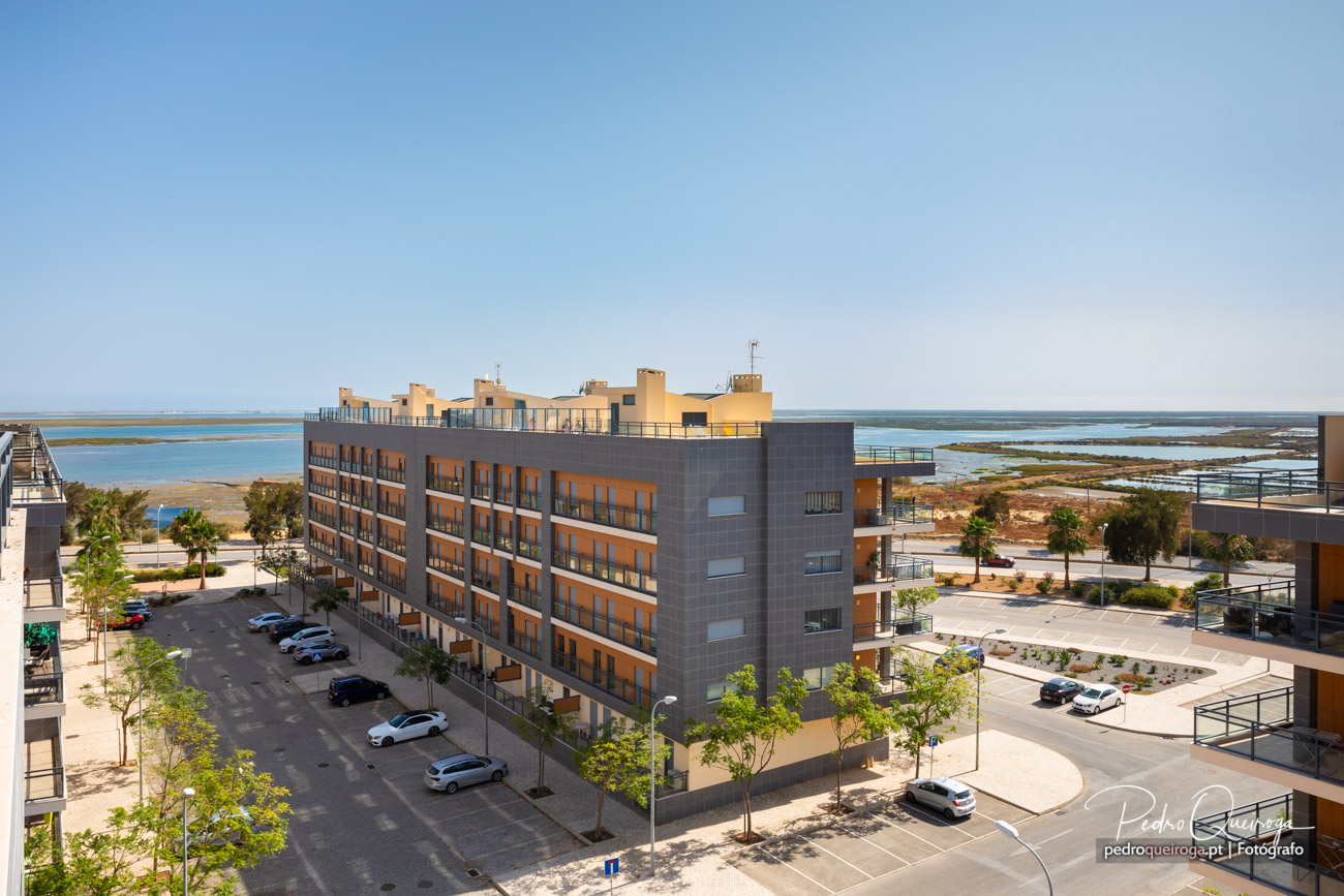 Edifício moderno com varandas laranja junto à Ria Formosa, rodeado por estacionamento e palmeiras sob céu azul.