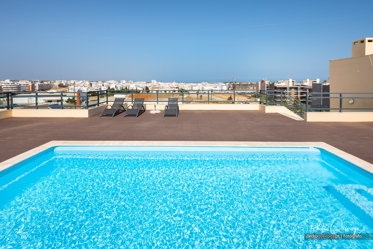 Piscina de águas cristalinas num rooftop amplo com espreguiçadeiras e vista panorâmica sobre a cidade ao fundo