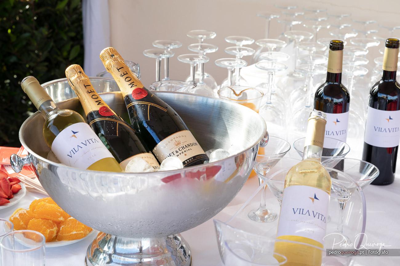 Garrafas de champanhe e vinho branco em balde de gelo prateado, rodeadas por taças de cristal e garrafas de vinho tinto