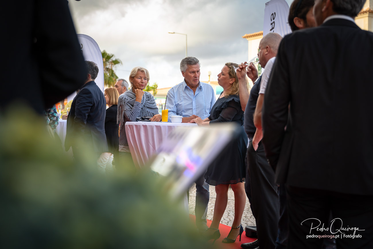 Convidados conversam animadamente em torno de mesa alta durante receção ao ar livre num dia nublado
