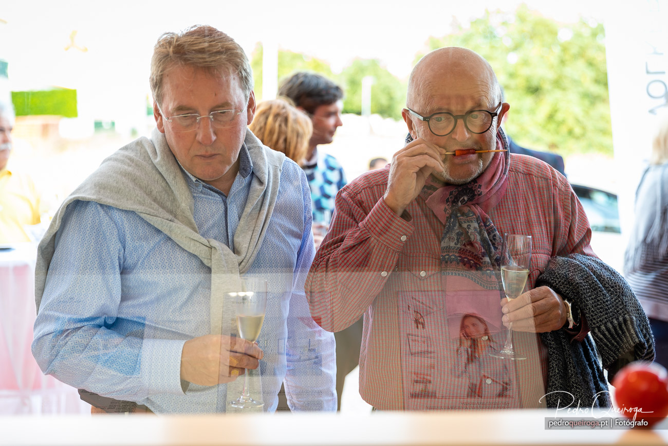 Dois homens provam vinho branco num evento social descontraído, rodeados por outros convidados em ambiente luminoso.
