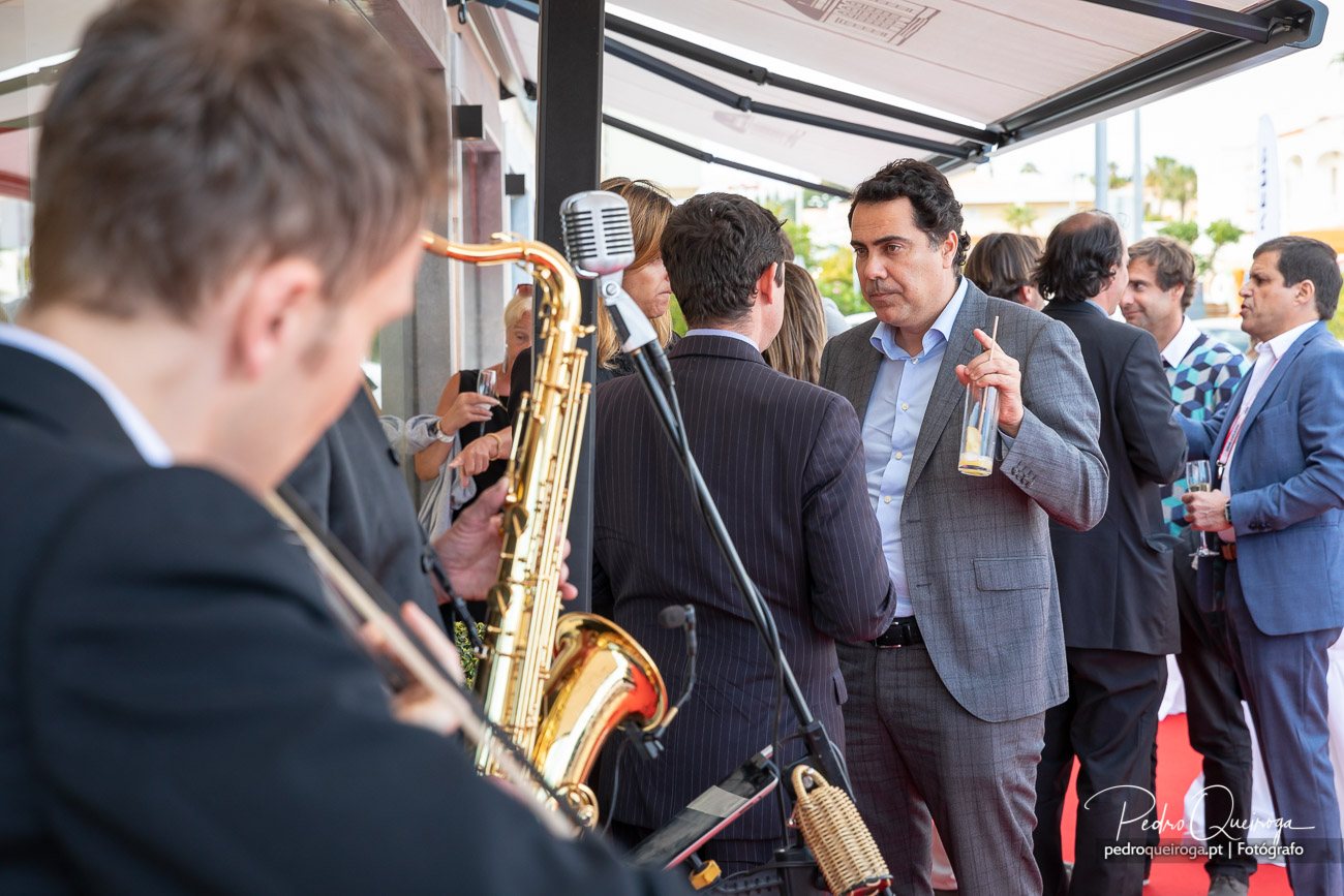 Convidados em fato conversam num evento ao ar livre enquanto músico toca saxofone dourado junto a microfone vintage