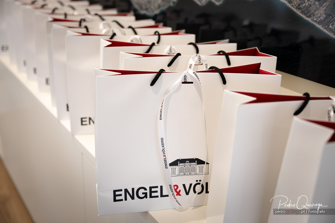 Sacos de papel brancos alinhados com interior vermelho e alças pretas sobre mesa clara em evento corporativo