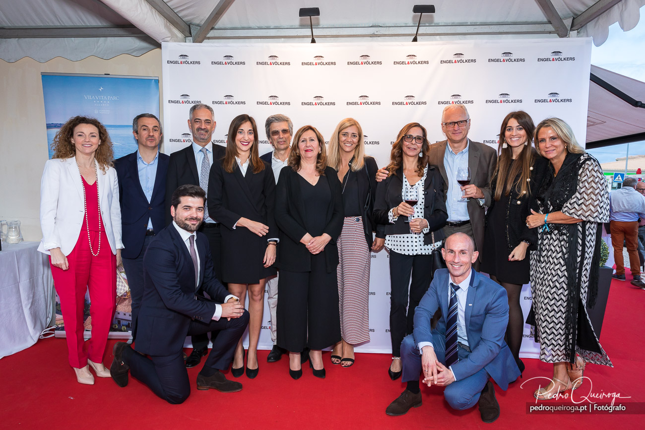 Equipa profissional posando em passadeira vermelha diante de painel corporativo num evento empresarial elegante.
