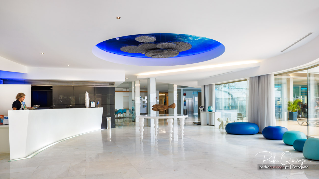 Hall de hotel moderno com receção branca, teto iluminado a azul e puffs coloridos