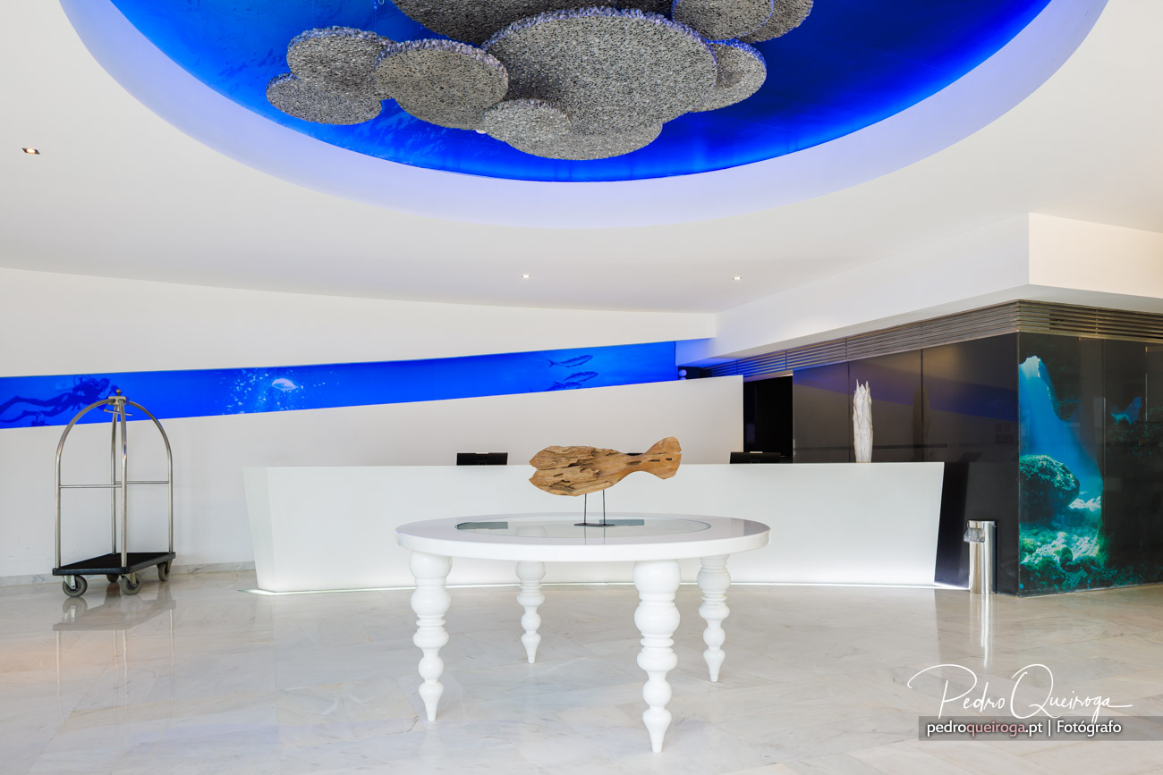 Hall de hotel moderno com teto azul vibrante, escultura em madeira sobre mesa branca e aquário iluminado ao fundo