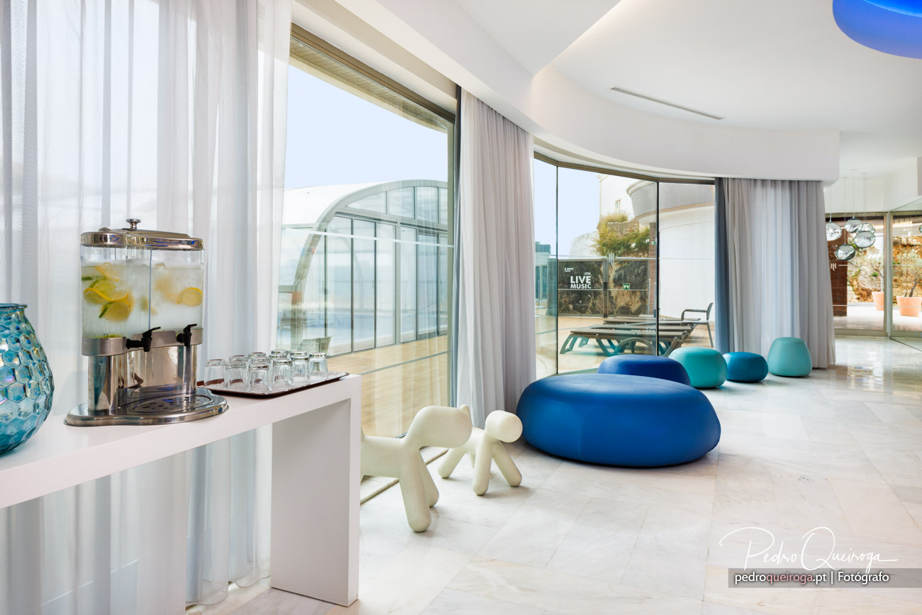 Sala moderna e luminosa com mobiliário azul e branco, amplas janelas e vista para terraço exterior com piscina