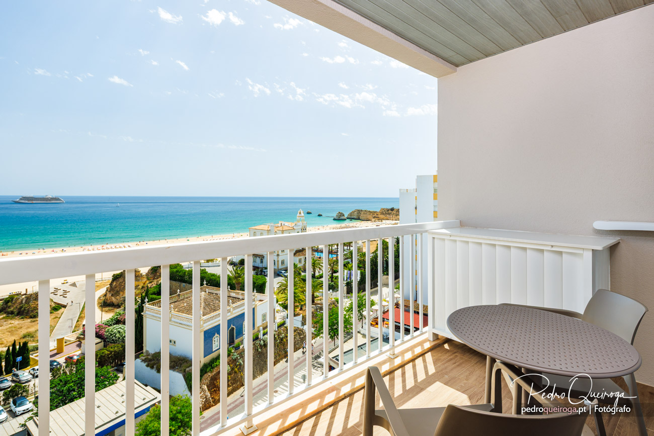 Varanda ampla com vista panorâmica sobre o mar azul-turquesa, praia dourada e navio cruzeiro ao longe no horizonte