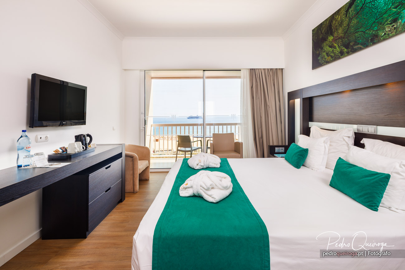 Quarto de hotel luminoso com vista mar, cama decorada em verde esmeralda e varanda privada com cadeiras