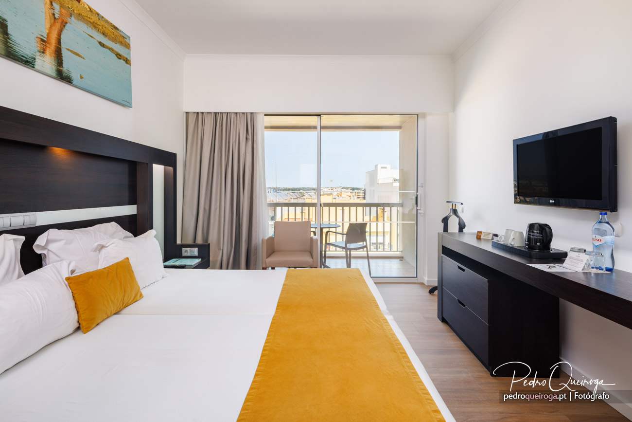 Quarto de hotel moderno com cama de casal, detalhes mostarda, varanda com vista urbana e televisão de parede