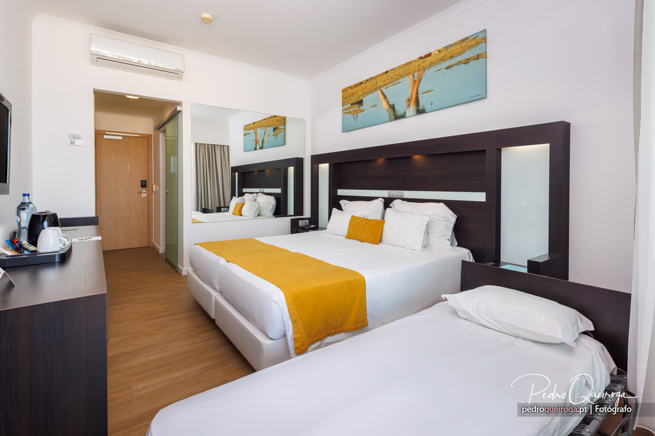 Quarto de hotel moderno com duas camas, cabeceira em madeira escura, manta amarela e quadros de praia na parede