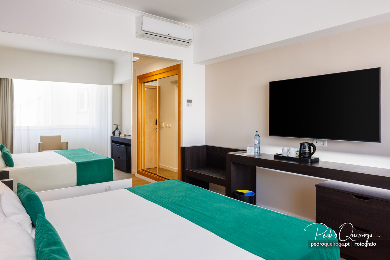 Quarto de hotel moderno com duas camas, mantas verdes, televisão de parede e ar condicionado instalado no teto branco.