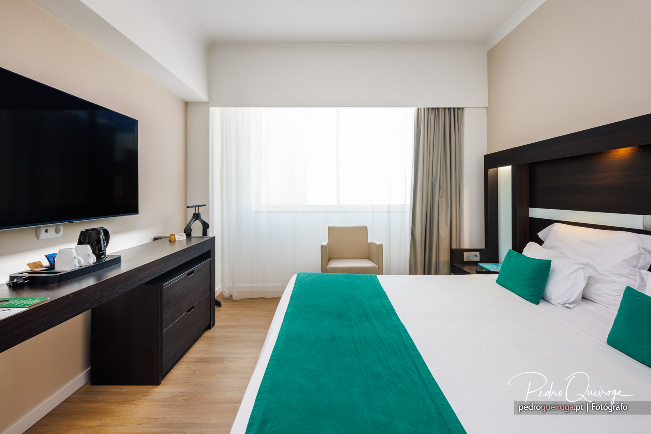 Quarto de hotel moderno com cama dupla, detalhes turquesa, televisão, poltrona bege e ampla janela com cortinas claras