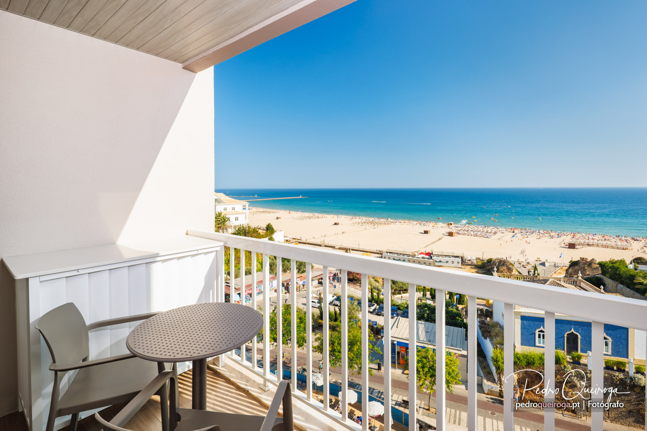 Varanda branca com mesa e cadeira, vista panorâmica sobre praia dourada e mar azul turquesa ao fundo.