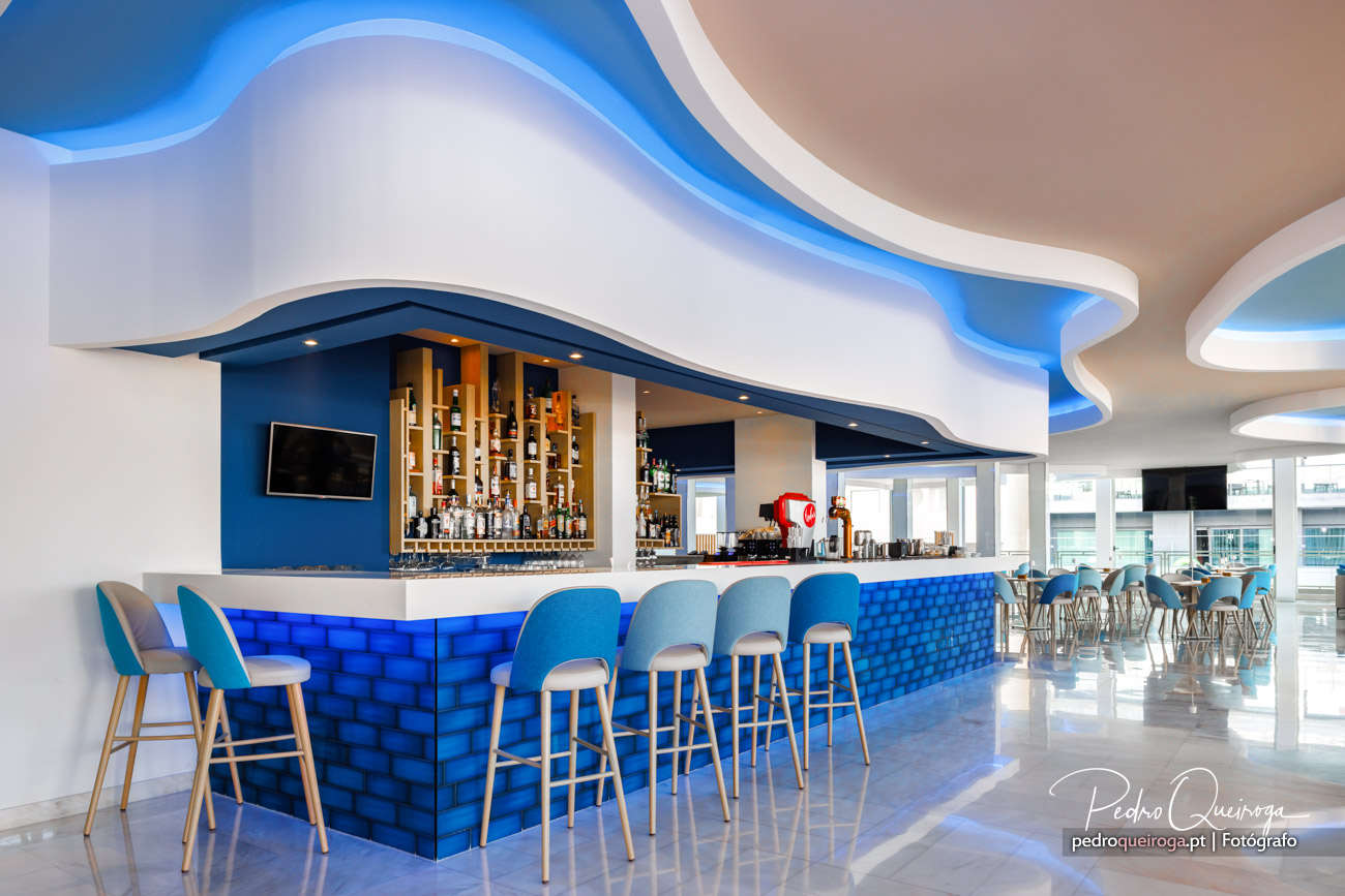 Bar moderno de hotel com bancada azul iluminada, cadeiras altas e teto ondulado com luz ambiente azul