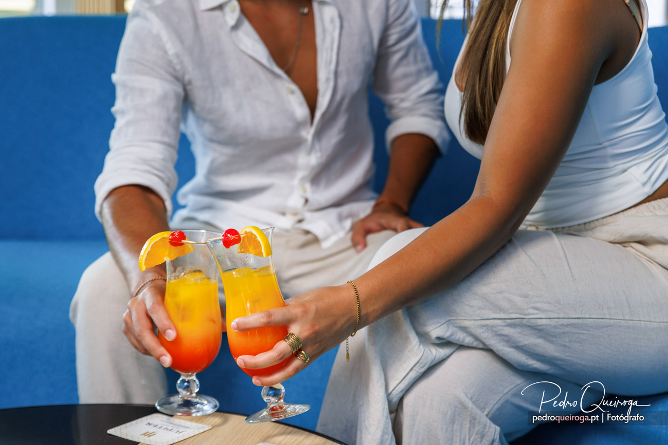 Casal sentado lado a lado segura cocktails coloridos decorados com laranja e cereja junto a parede azul vibrante