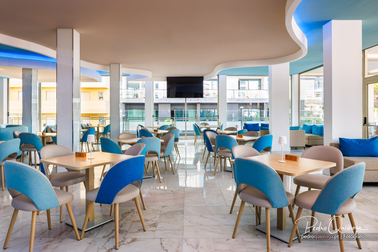 Sala de refeições moderna com cadeiras azuis e bege, teto ondulado e amplas janelas com luz natural abundante