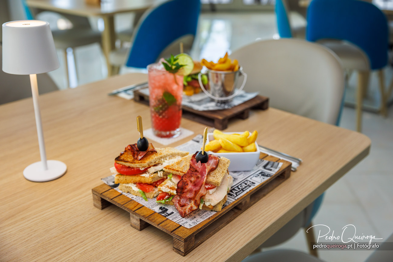 Sandes club com bacon crocante acompanhadas de batatas fritas, cocktail colorido e snacks num ambiente moderno e acolhedor