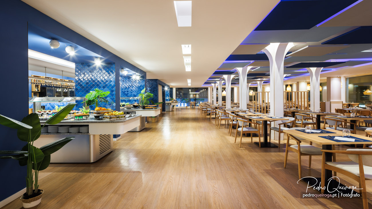 Restaurante de hotel moderno com buffet azulejado, colunas brancas orgânicas e iluminação LED ambiente azul no teto
