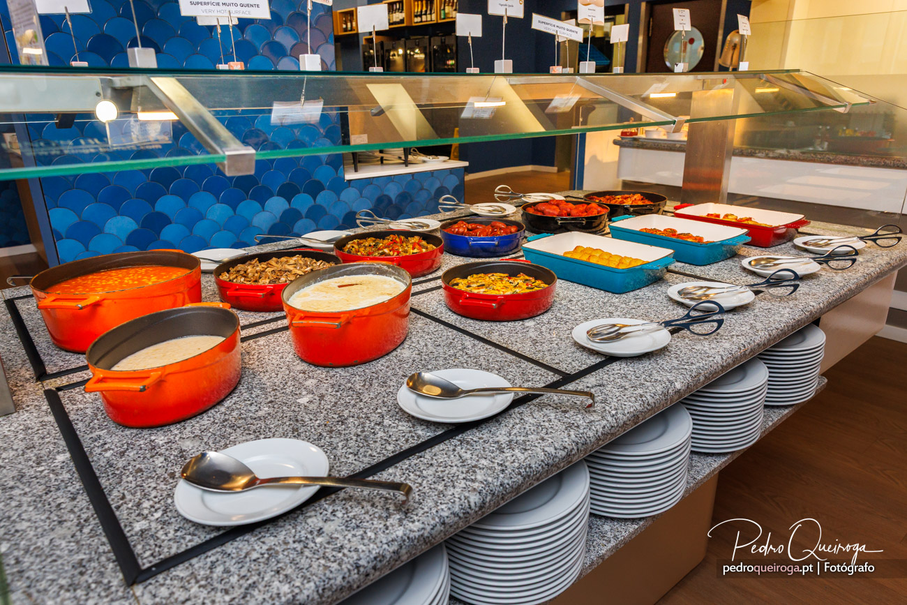 Buffet de hotel com variedade de pratos quentes em recipientes coloridos sobre bancada de granito com pratos empilhados