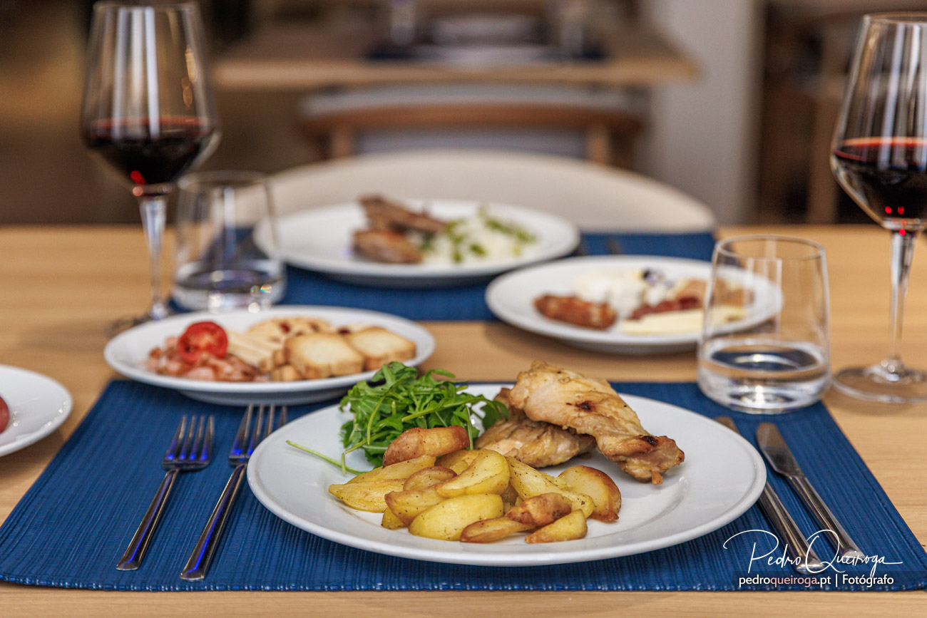 Mesa de restaurante com pratos de frango grelhado, batatas assadas e rúcula, acompanhados por copos de vinho tinto e água.