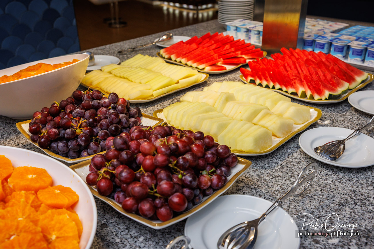 Mesa de buffet com travessas de uvas vermelhas, queijo fatiado, melancia e outros frutos dispostos em bancada de granito