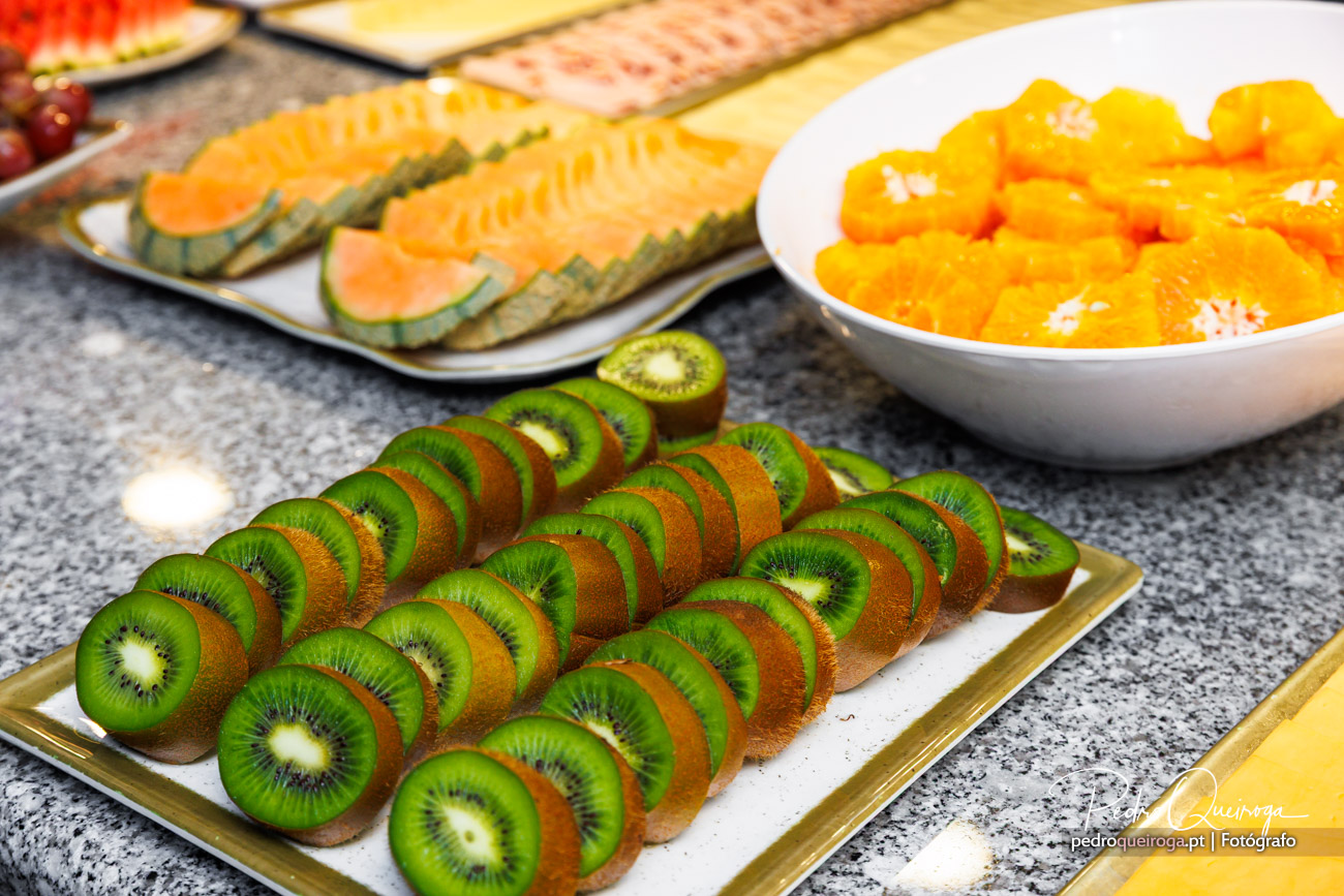 Rodelas de kiwi dispostas em bandeja dourada, com melão em taça branca e fatias de melancia ao fundo numa mesa de buffet