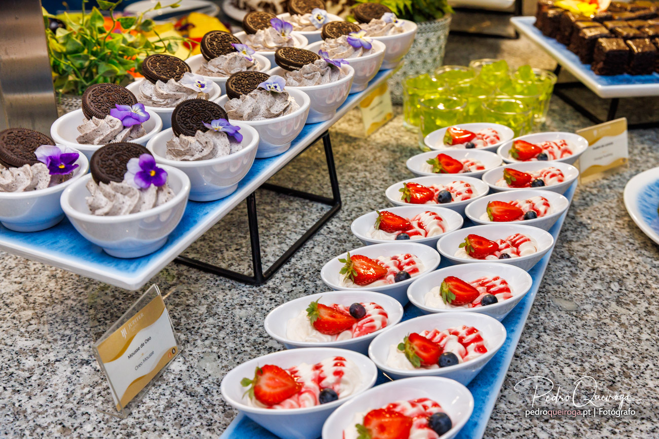 Filas de sobremesas em taças brancas: mousse de chocolate com bolachas e violetas, panna cotta com frutos vermelhos e gelatina verde ao fundo