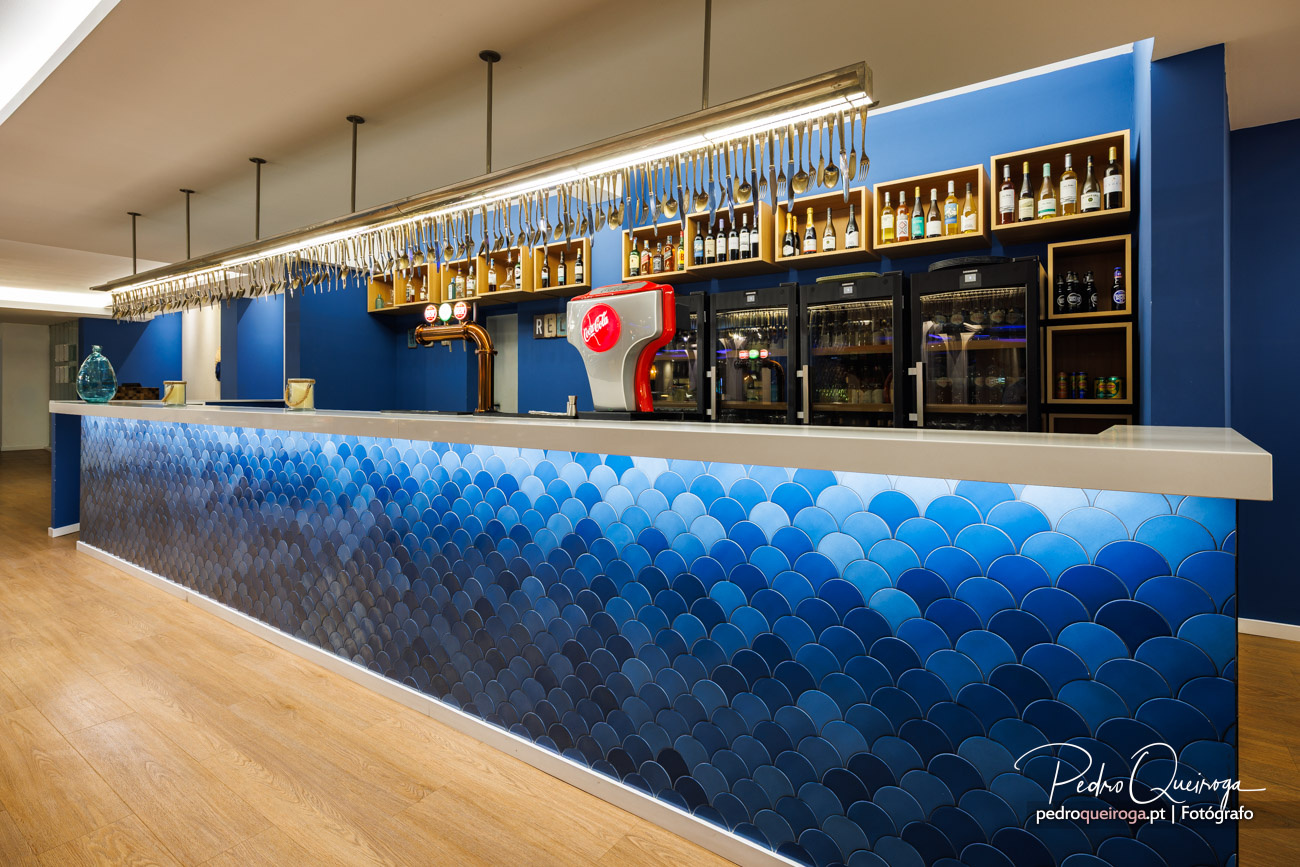 Bar moderno com balcão revestido a azulejos azuis em escama, prateleiras iluminadas e máquina de cerveja vermelha