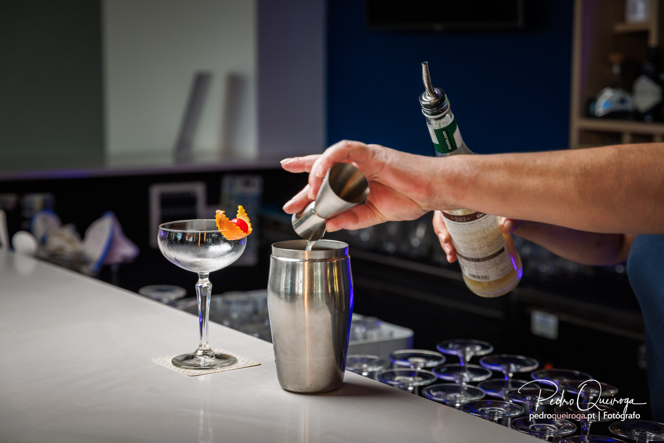 Barman a preparar cocktail com shaker em balcão branco, taça com decoração de casca de laranja ao lado