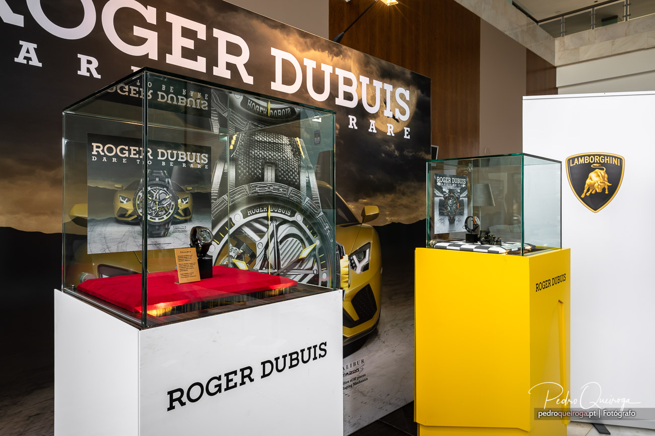 Exposição de relógios Roger Dubuis em vitrines elegantes com Lamborghini amarelo ao fundo num showroom moderno
