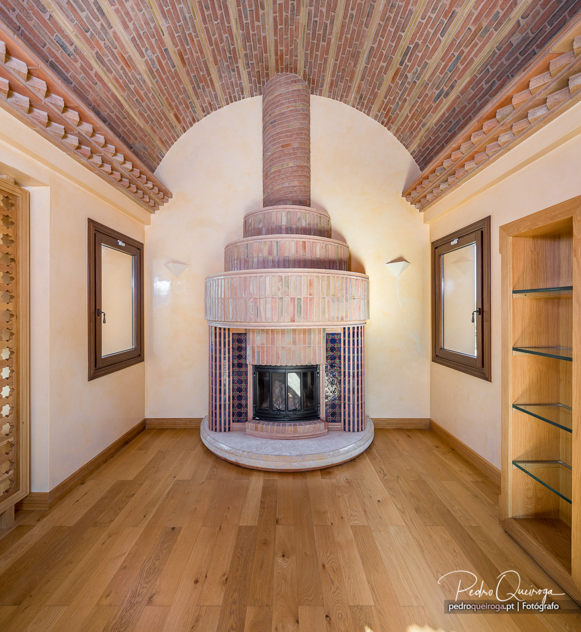 Lareira cilíndrica em tijolo com azulejos decorativos sob teto abobadado em tijolo aparente e chão de madeira clara