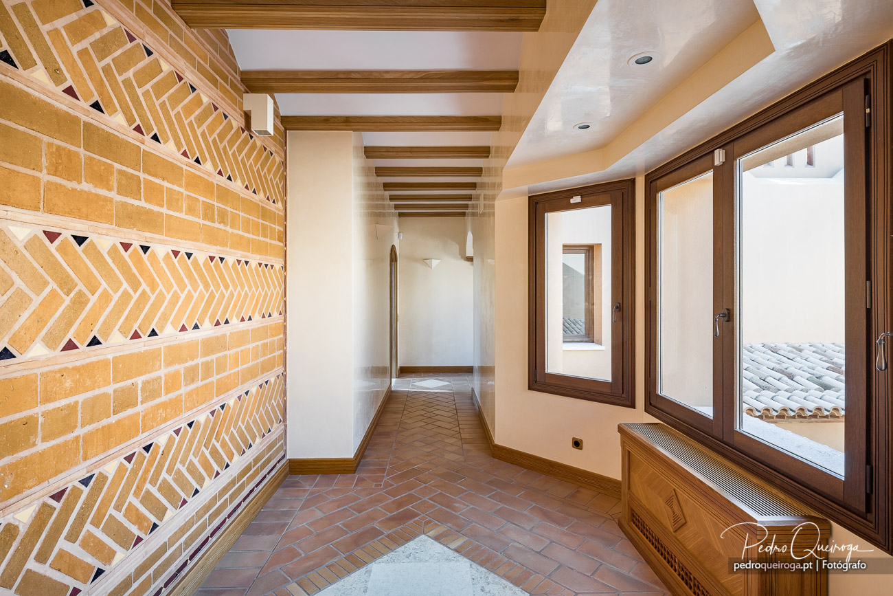 Corredor amplo com parede de tijolo amarelo decorativo, vigas de madeira no teto e janelas com vista para telhados