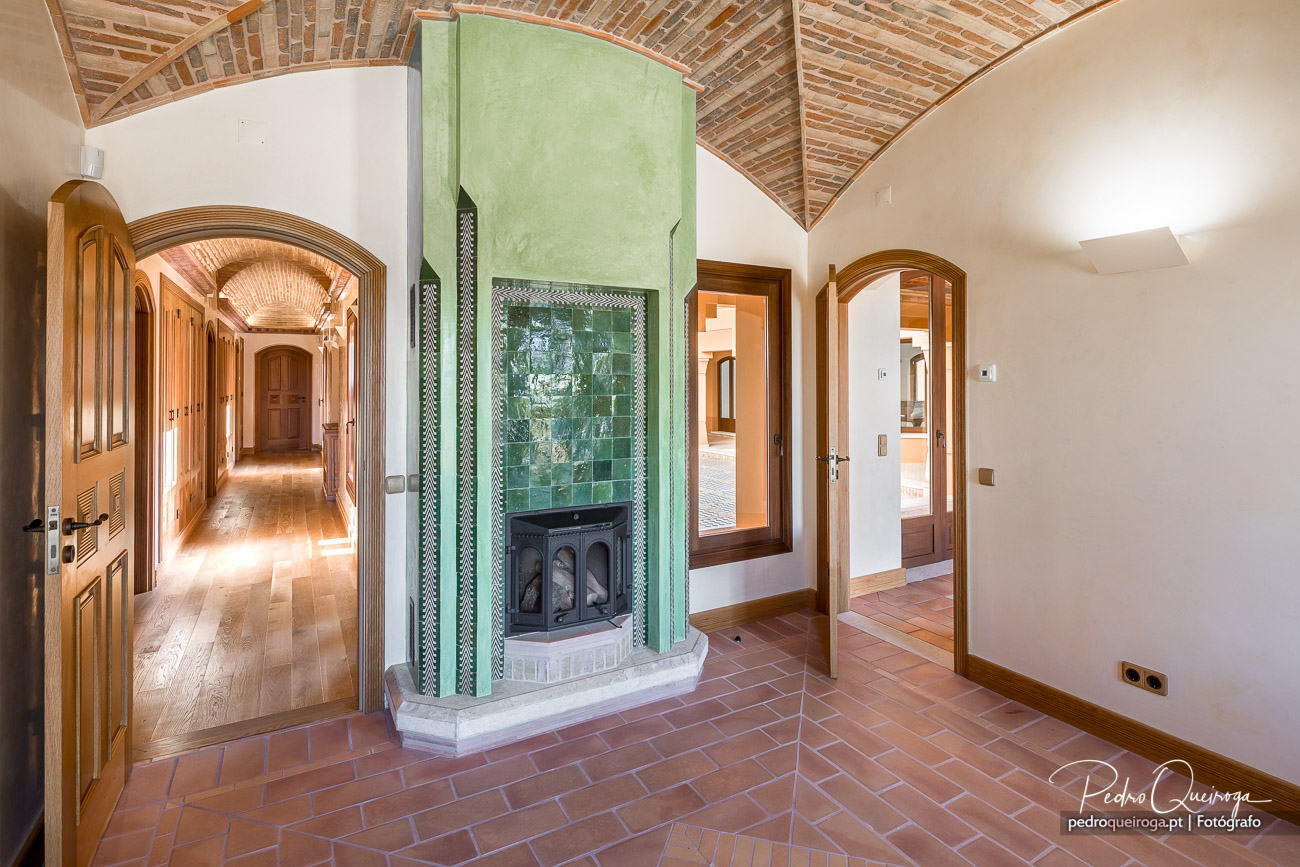 Hall elegante de hotel com lareira verde em azulejo, tetos abobadados em tijolo e corredor iluminado ao fundo