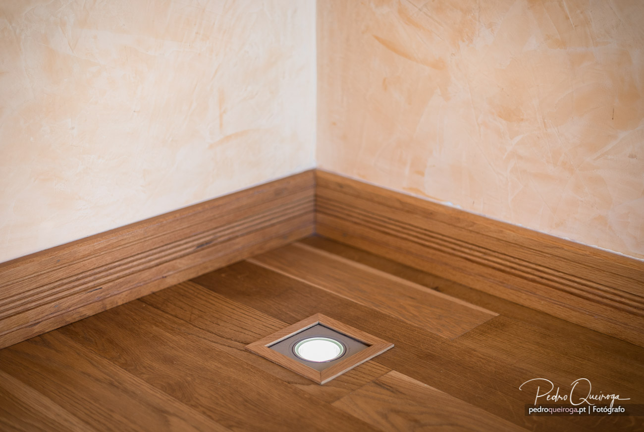 Detalhe de rodapé em madeira com foco embutido no chão, junto a parede clara com textura suave