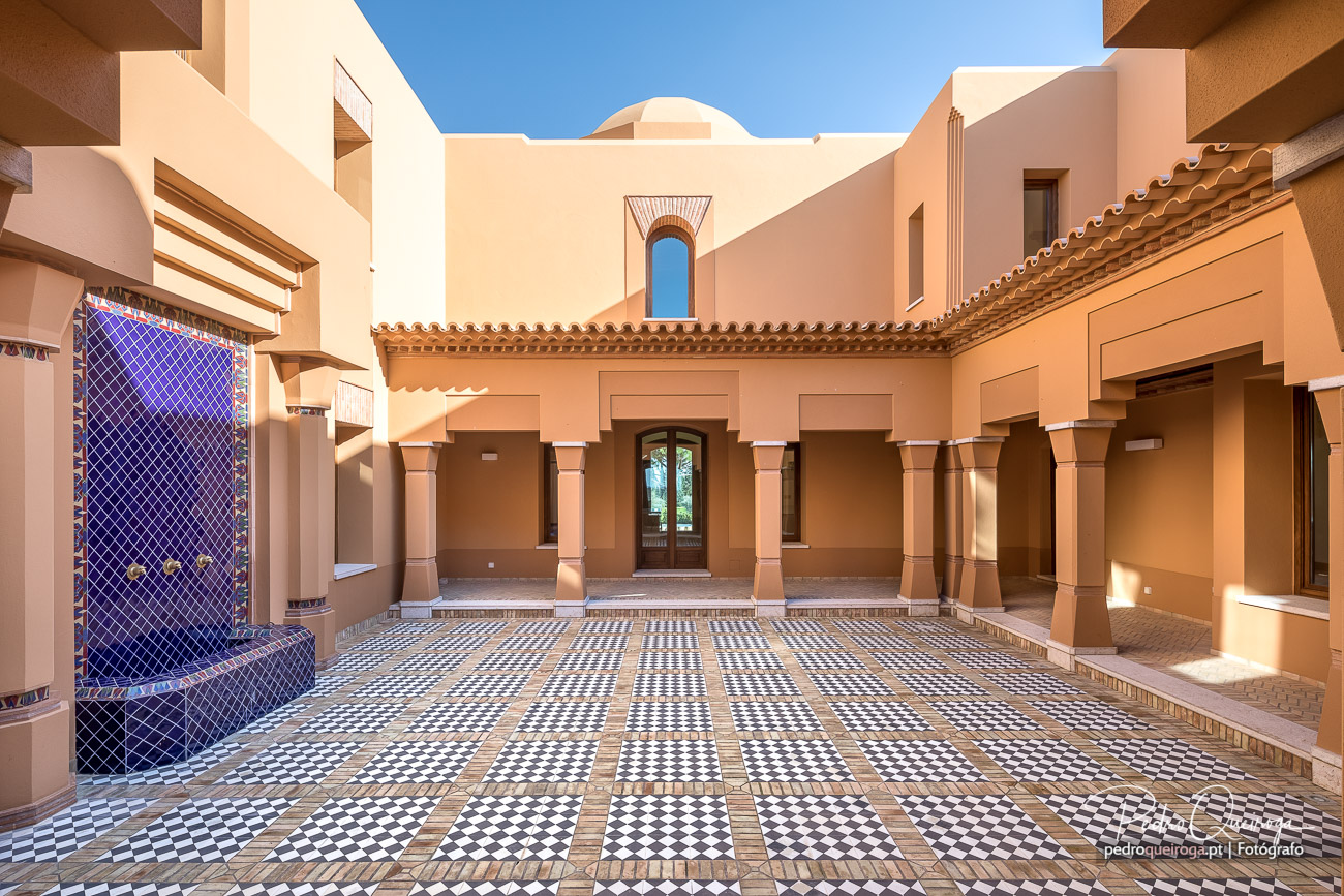 Pátio interior com arcos em tons terrosos, mosaico geométrico no chão e fonte azul decorativa à esquerda sob céu azul
