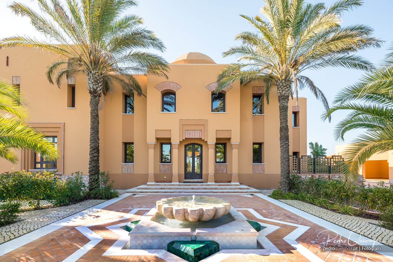 Moradia em tons de areia com palmeiras e fonte central, numa composição fotográfica arquitetónica elegante e luminosa.